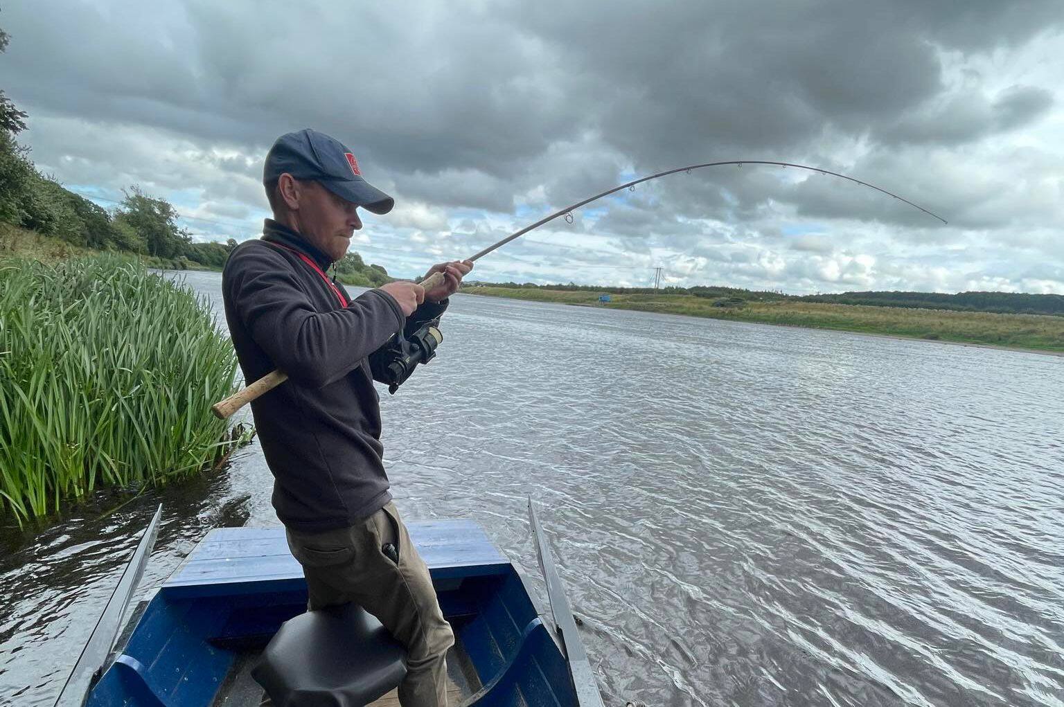 An angler standing in the boat with his rod bent into a fish on a cloudy summer day