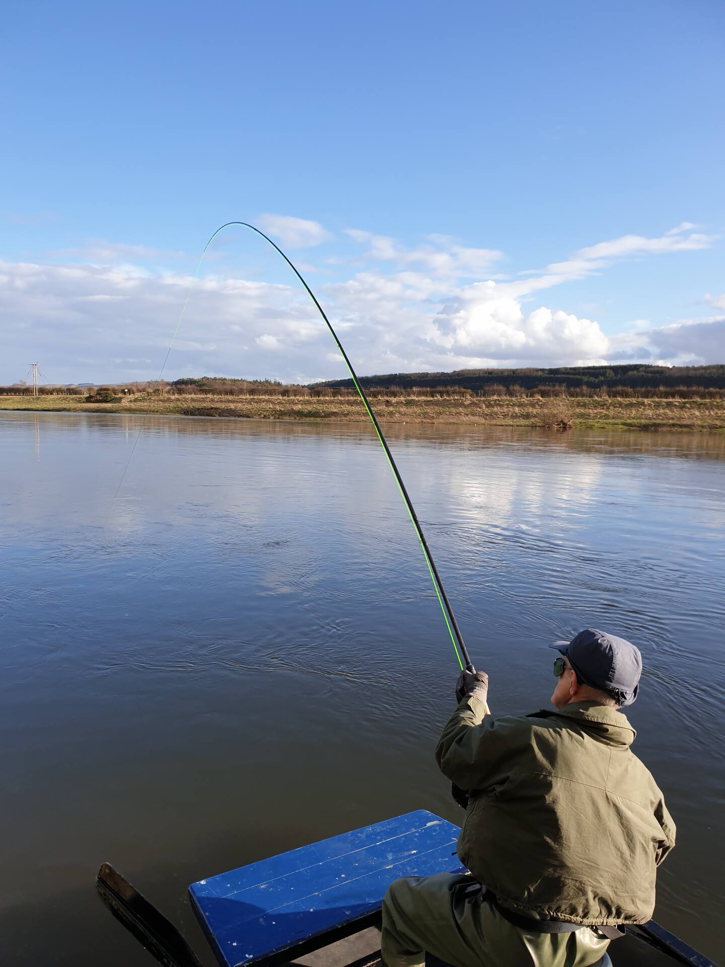 An angler in a boat with his rod bent into a fish on a bright day on the River Tweed