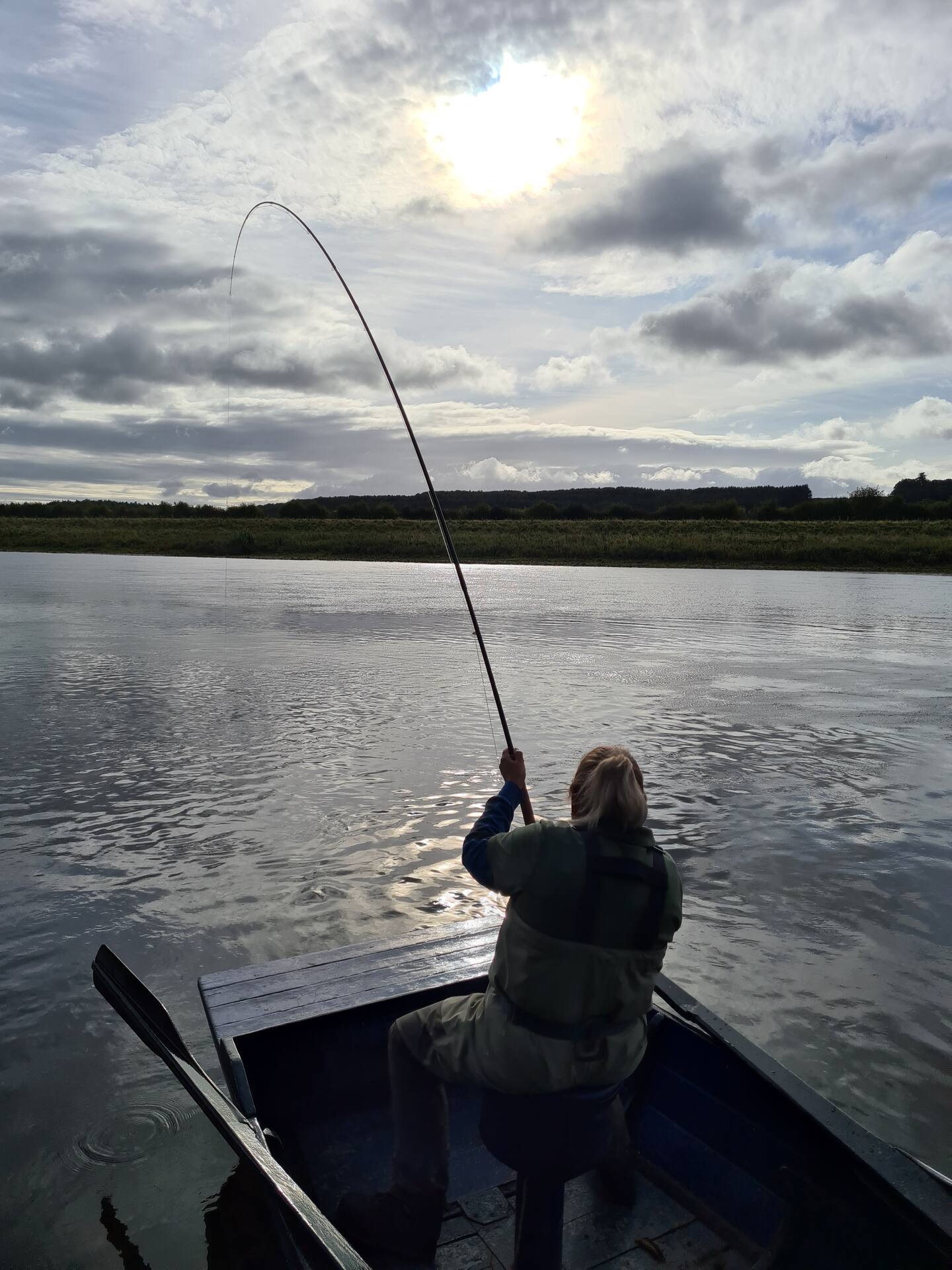 An angler in a boat playing a fish, rod bent, under an evening sky on the Tweed