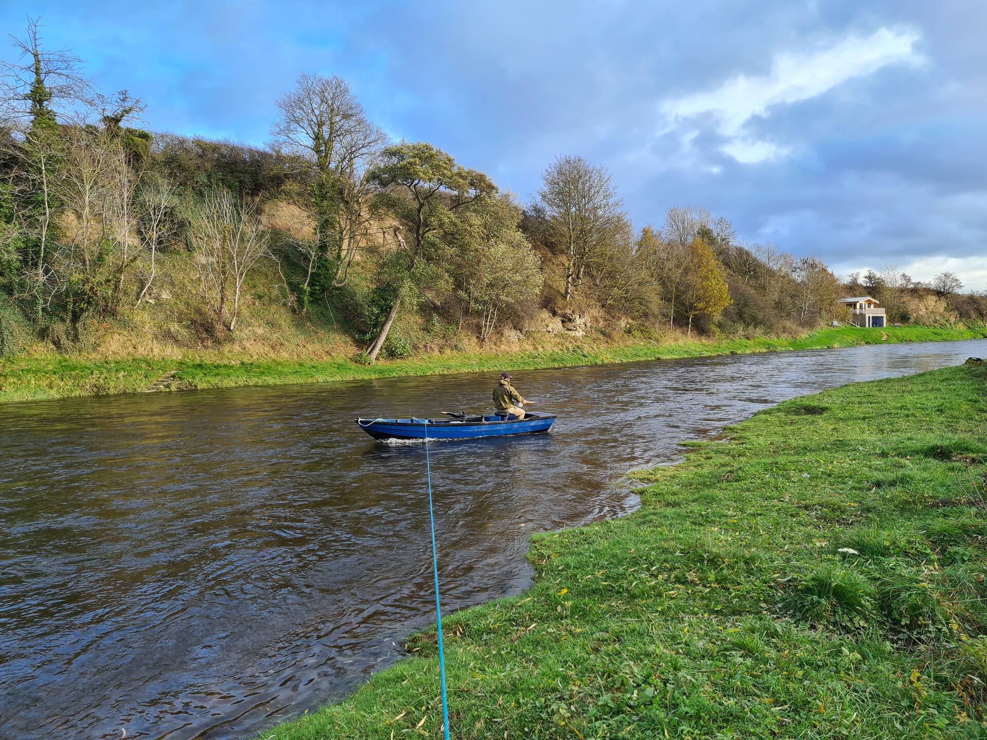 A blue boat with an angler on the Tweed below the wooded bank, the fishing hut in the distance