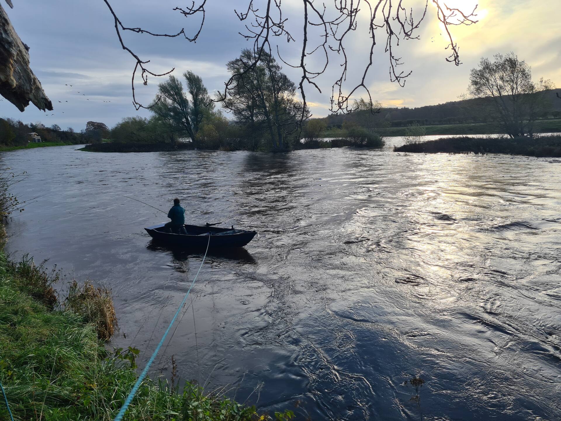 An angler fishing from a boat held on a rope from the bank, late autumn light