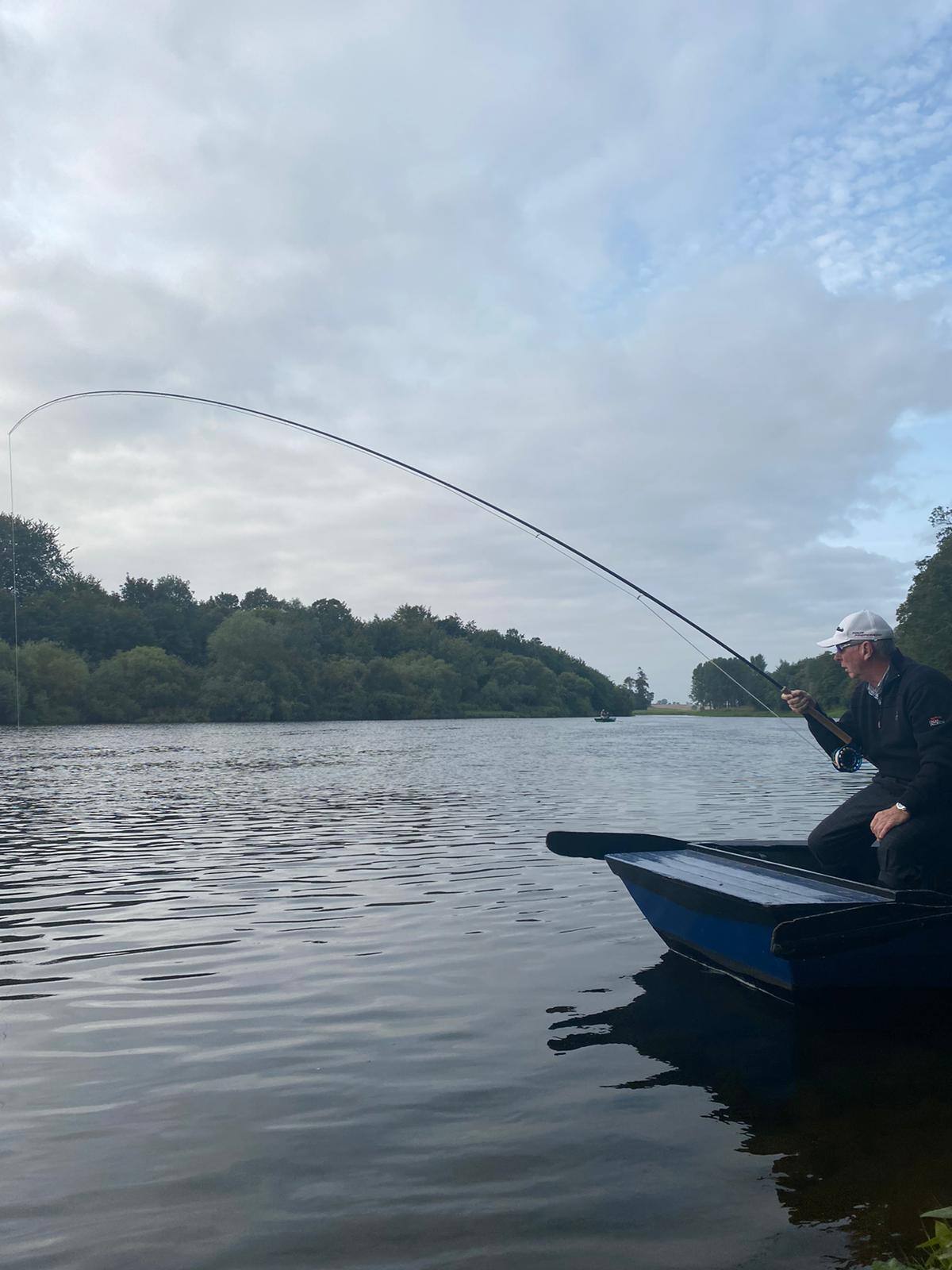 An angler fishing from the boat with his rod bent into a fish on a calm summer day
