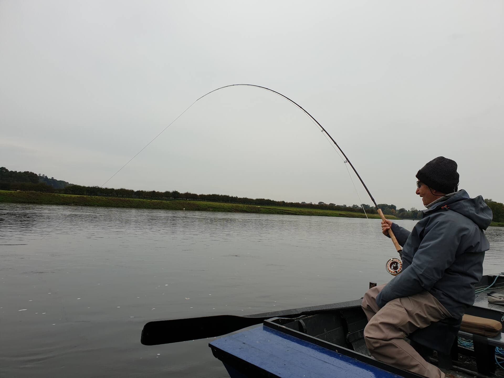 An angler playing a fish from a boat on an overcast day on the Tweed