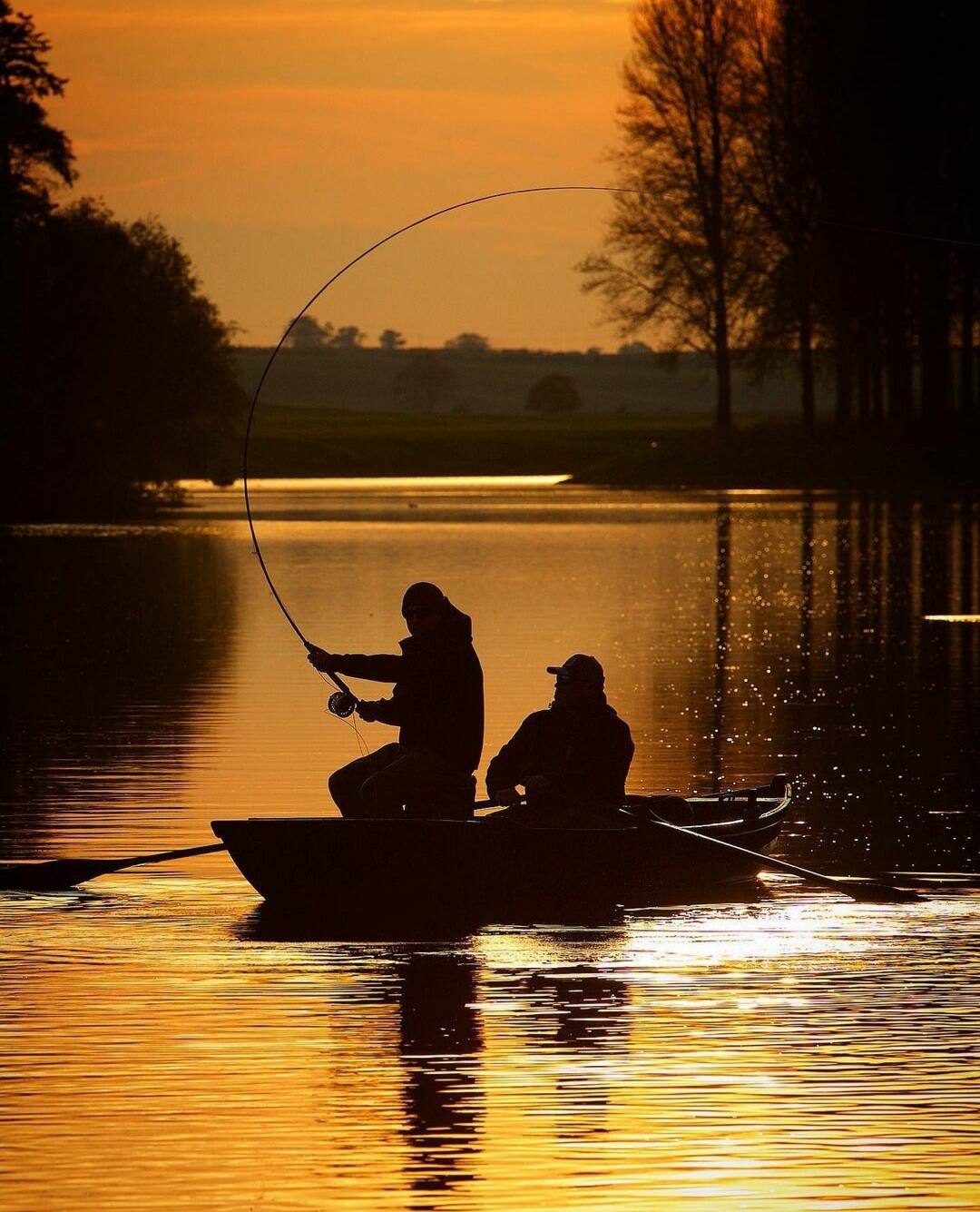Two anglers silhouetted in a boat at sunset, rod bent into a fish