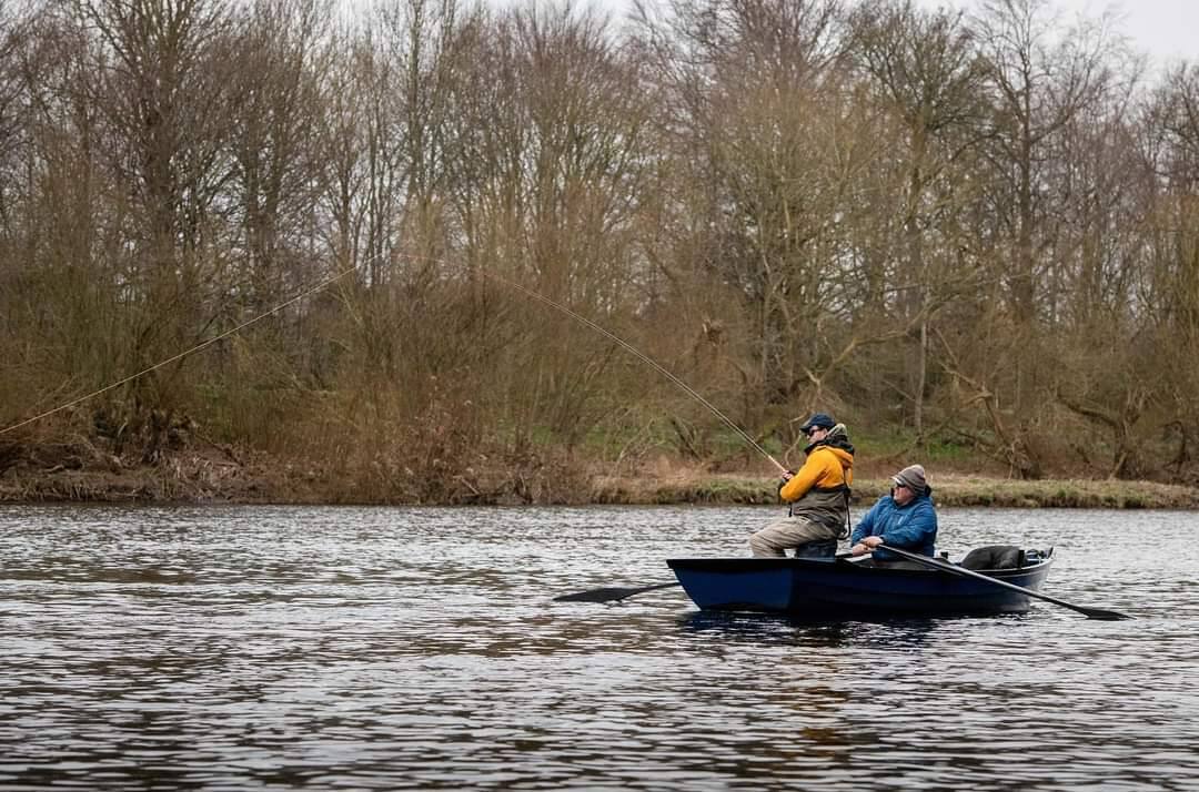 Two anglers in a blue boat fishing below bare spring trees on the Tweed