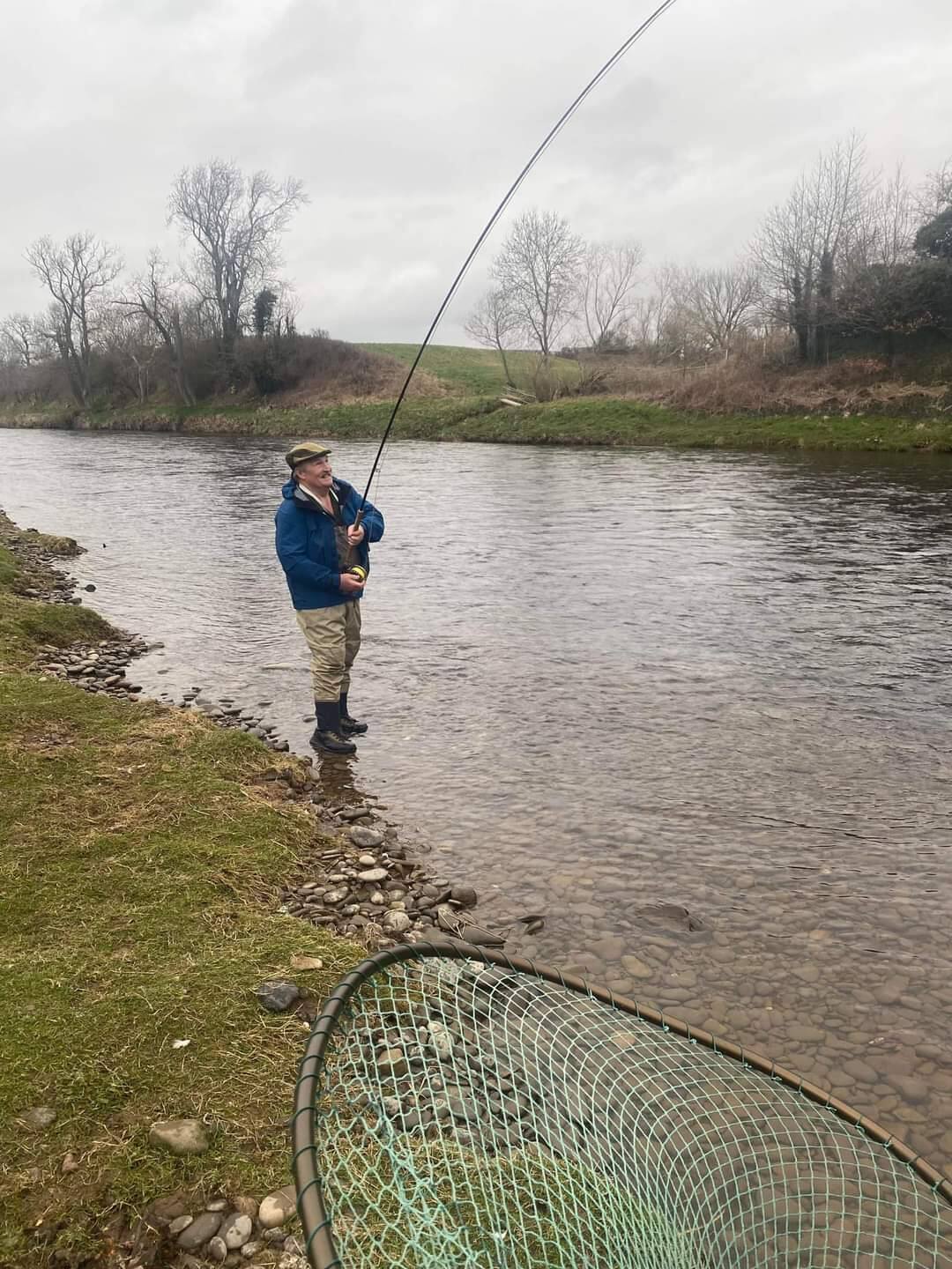 A wading angler playing a fish in the shallows, landing net ready on the bank